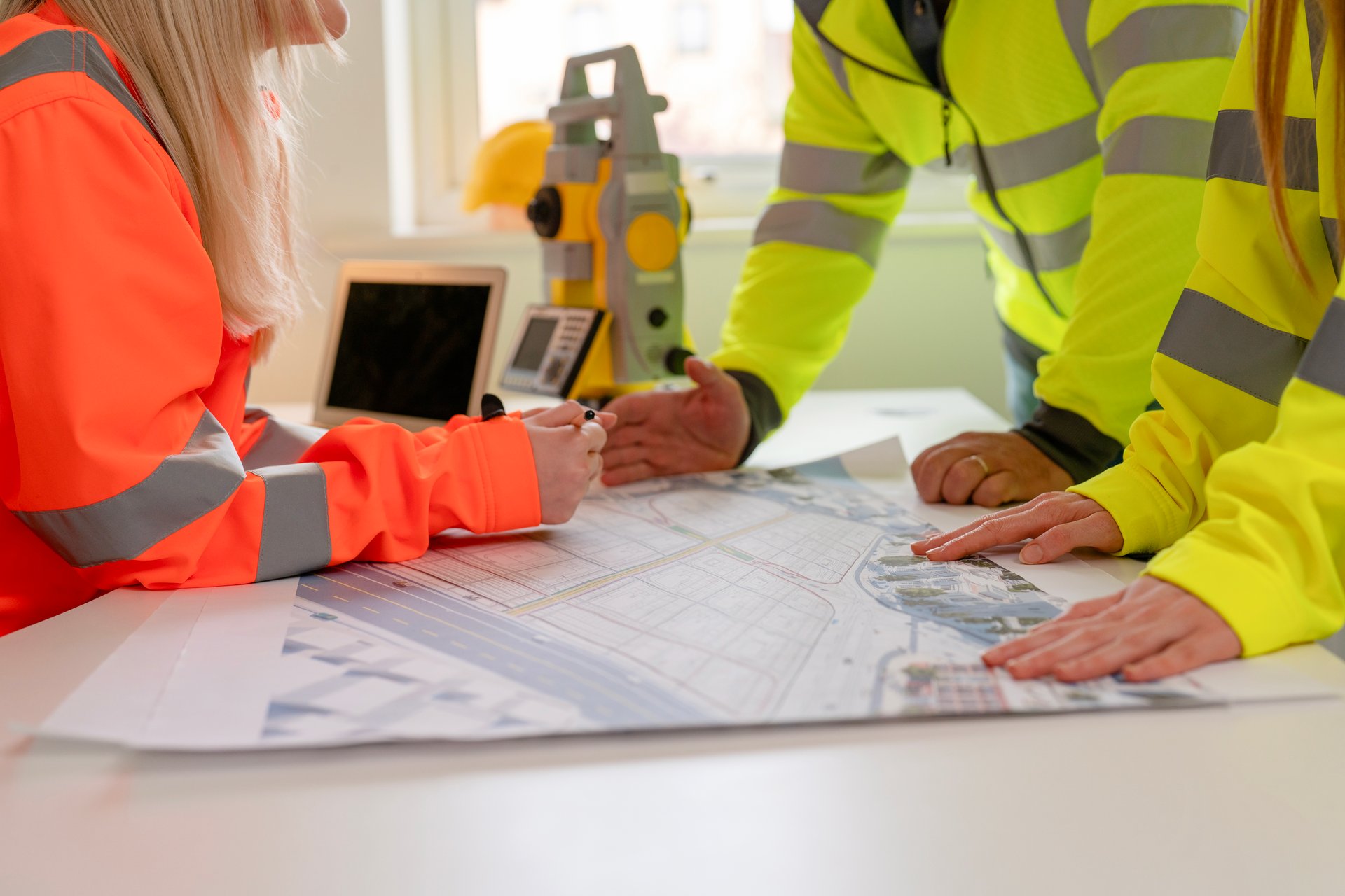 Engineering professionals collaborate on construction plans in bright office wearing safety hi-viz gear