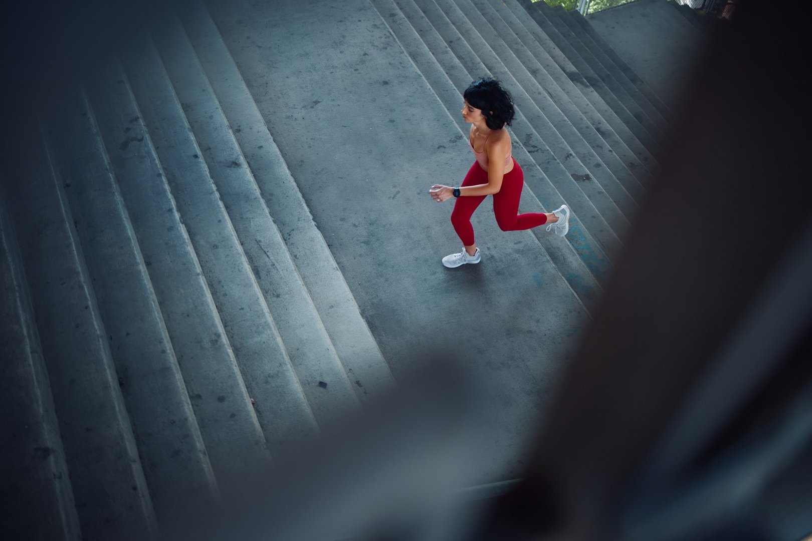Energetic woman wearing red activewear exercising by running up concrete stairs in an urban environment, representing fitness, health, and determination. A dynamic scene emphasizing movement and urban fitness lifestyle.