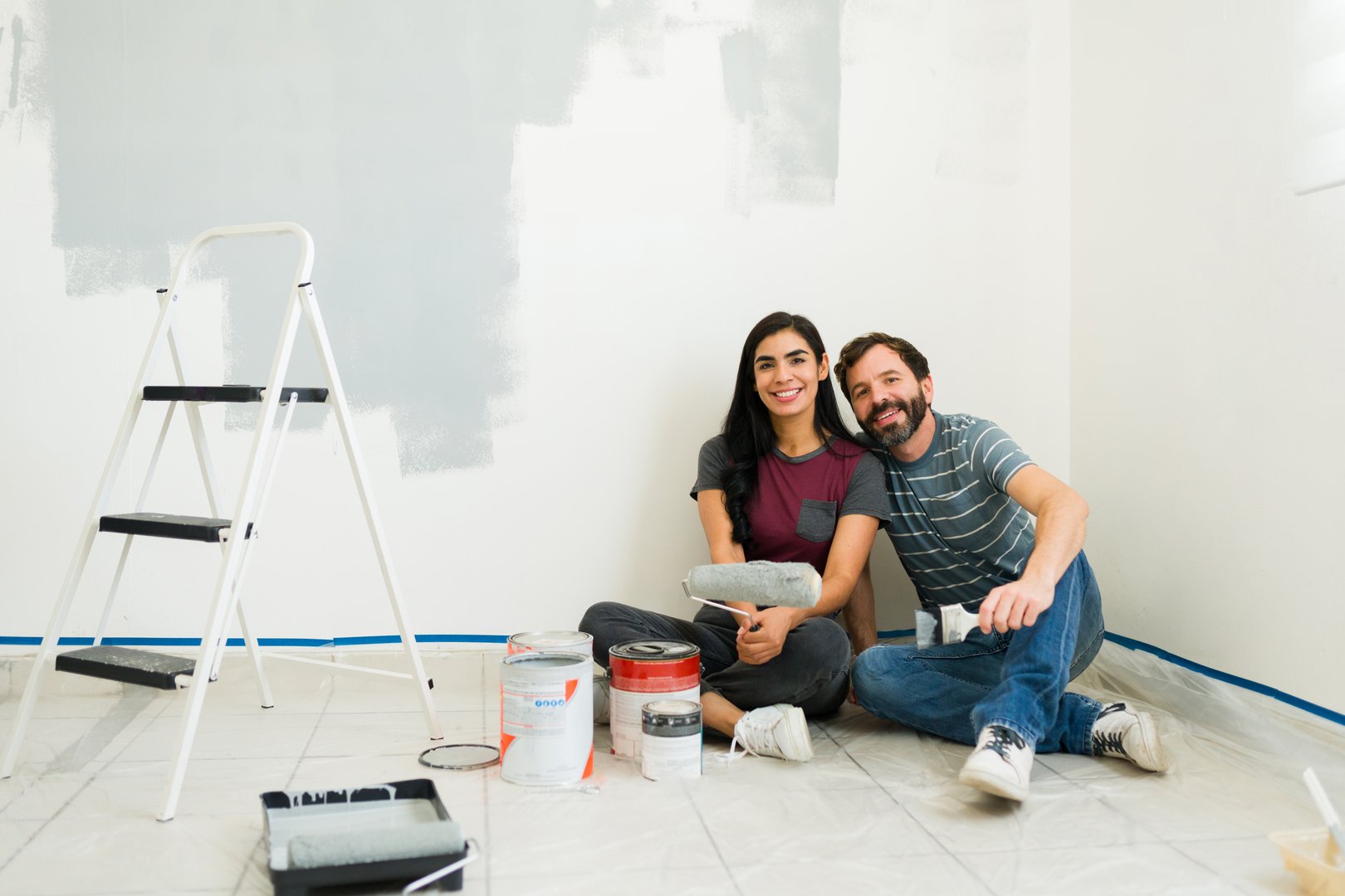 Young Hispanic couple taking a break while painting the walls gray in their new house, they are sitting on the floor and smiling