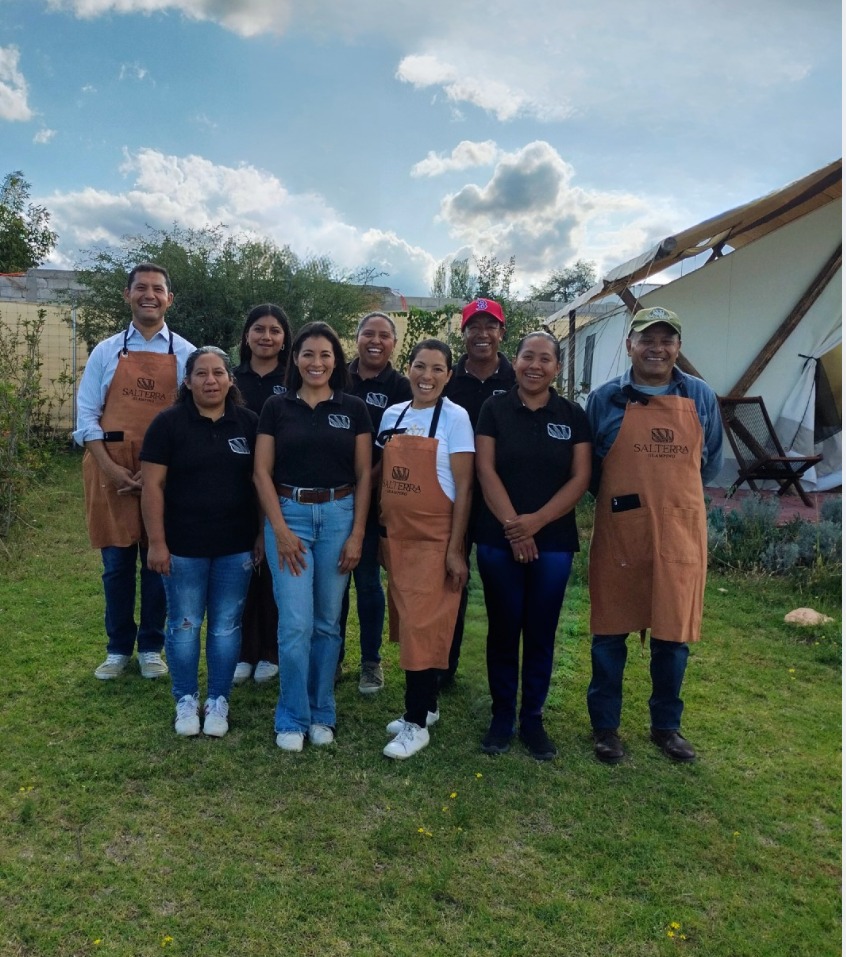 Group of smiling people wearing aprons, standing on grass with tents and trees in the background, under a partly cloudy sky.