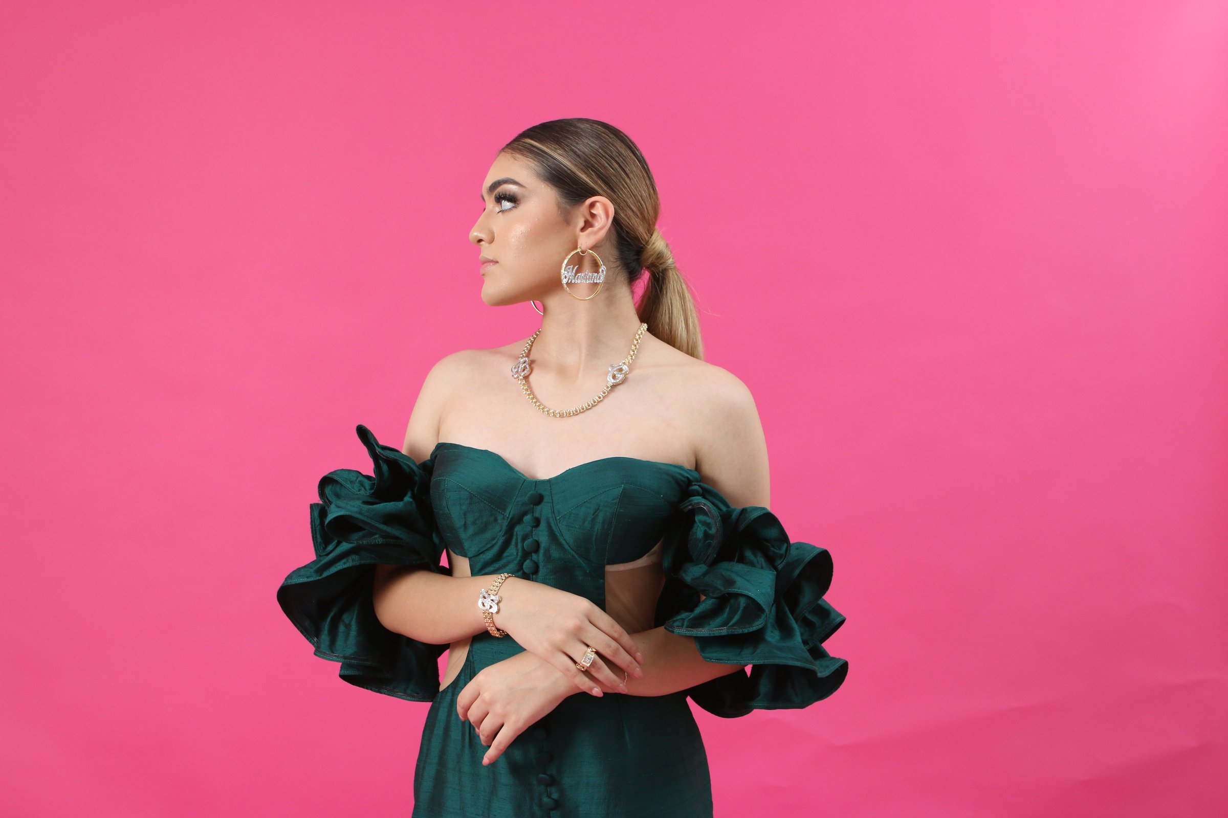 Woman in a green off-shoulder dress with ruffled sleeves, wearing jewelry, poses against a pink background.