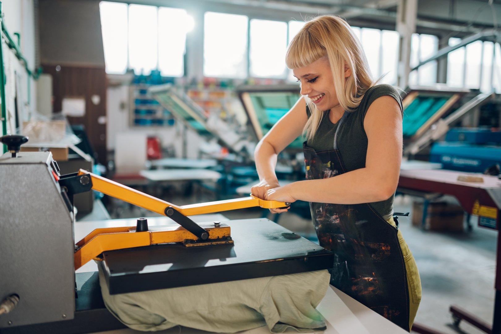 Happy young female graphic technology expert pressing heating press on textile product at printing workshop. Smiling printing workshop female employee using heat press on t-shirt. Females in industry.
