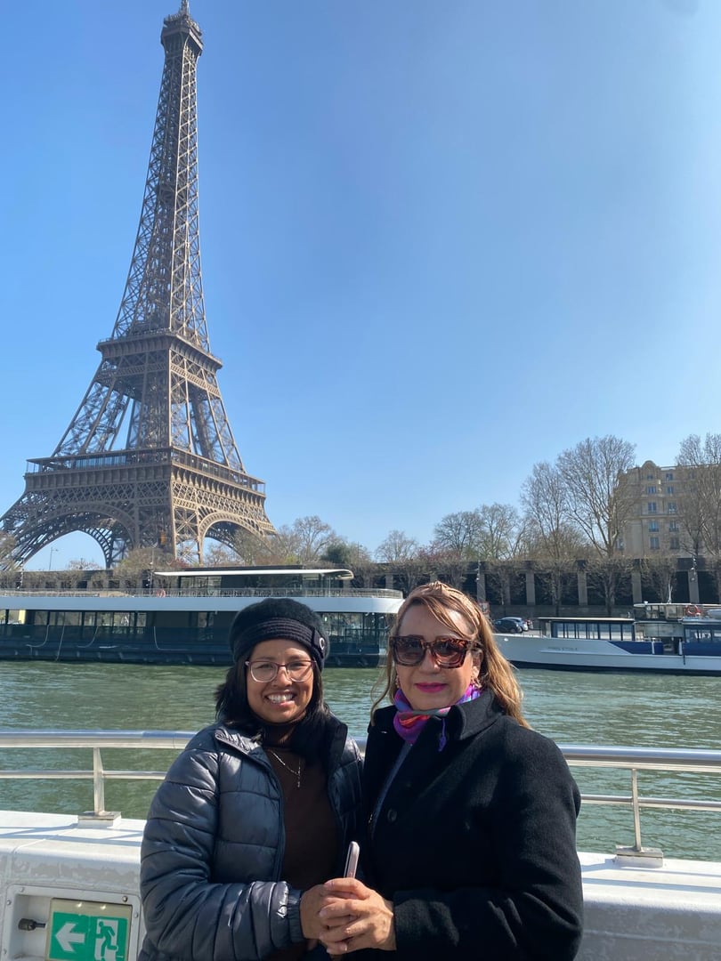 Two people smiling in front of the Eiffel Tower on a sunny day, standing by the Seine River with trees in the background.