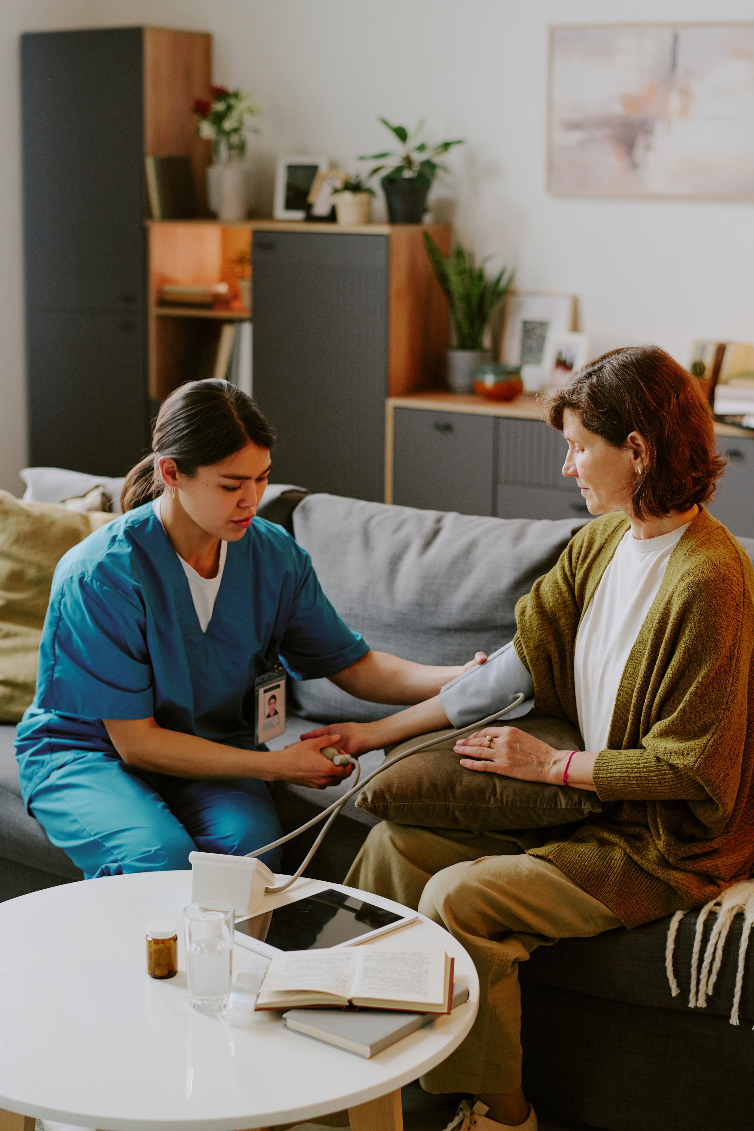 Elderly woman receiving blood pressure check from nurse in a living room setting, medical equipment and modern furniture visible in the background