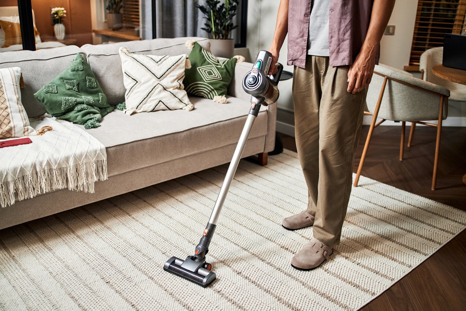 Man vacuuming carpet with cordless vacuum cleaner in living room, standing near sofa with decorative pillows, only lower body and hands visible