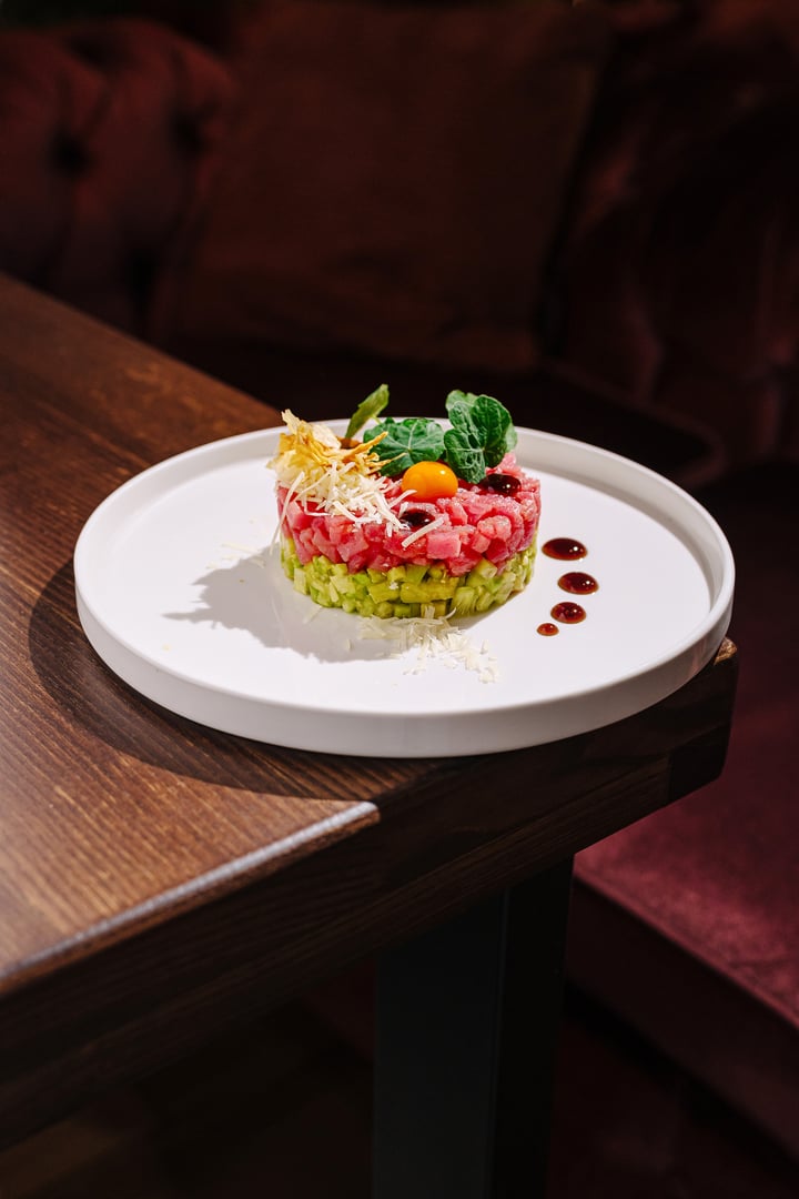 A beautifully crafted dish showcases layers of fresh vegetables and proteins, adorned with herbs and sauces. The plate is elegantly set on a wooden table, creating a warm dining atmosphere.