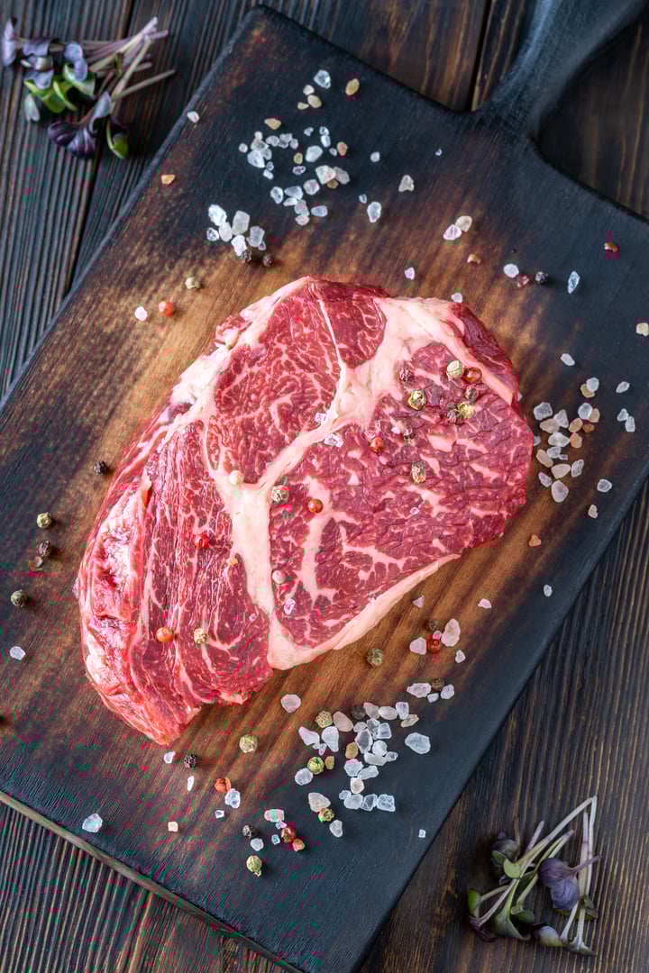 Thick, marbled ribeye steak resting on a rustic board, seasoned with salt and pepper