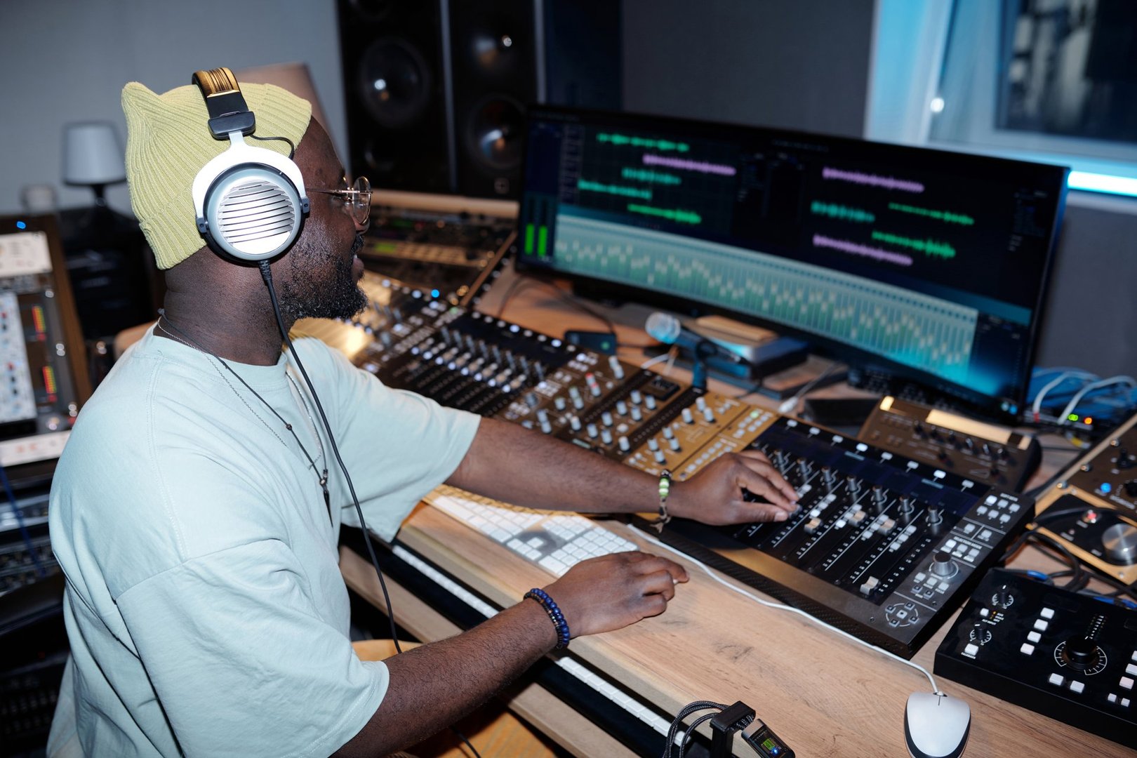 Music producer wearing headphones and a beanie carefully adjusting audio levels on a mixing console in modern recording studio environment filled with equipment and monitors