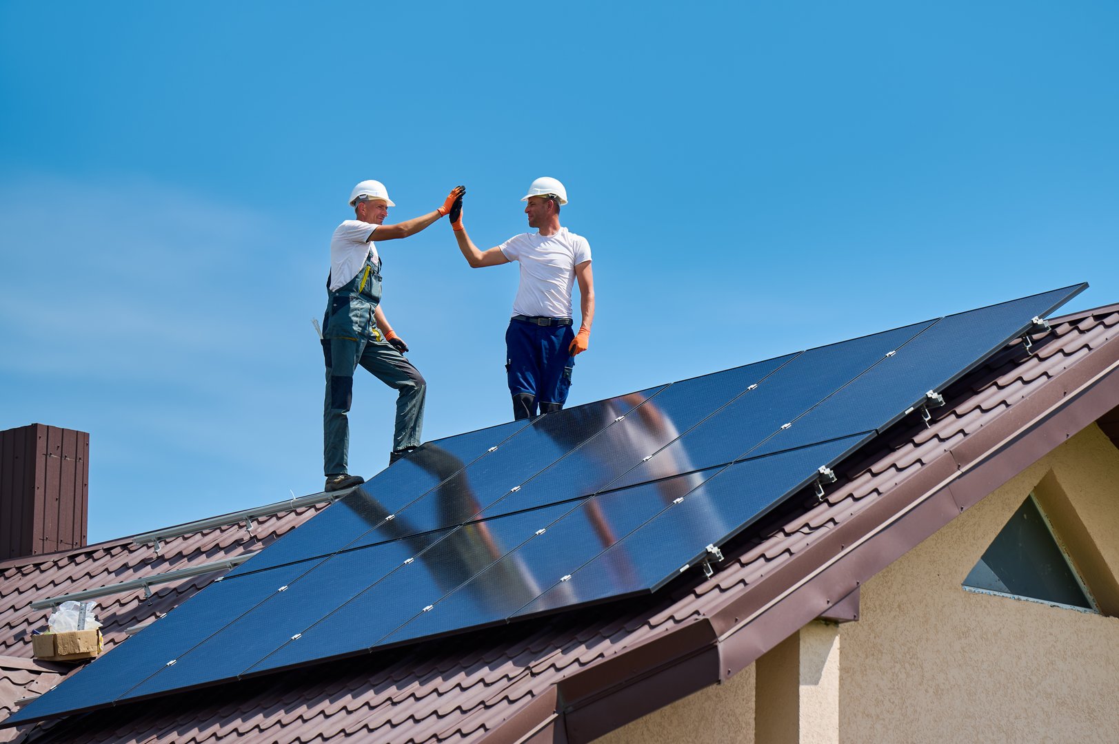Worker building solar panel system on metal roof of house. Engineers giving high five to each other, standing on rooftop while installing photovoltaic solar module outdoors.