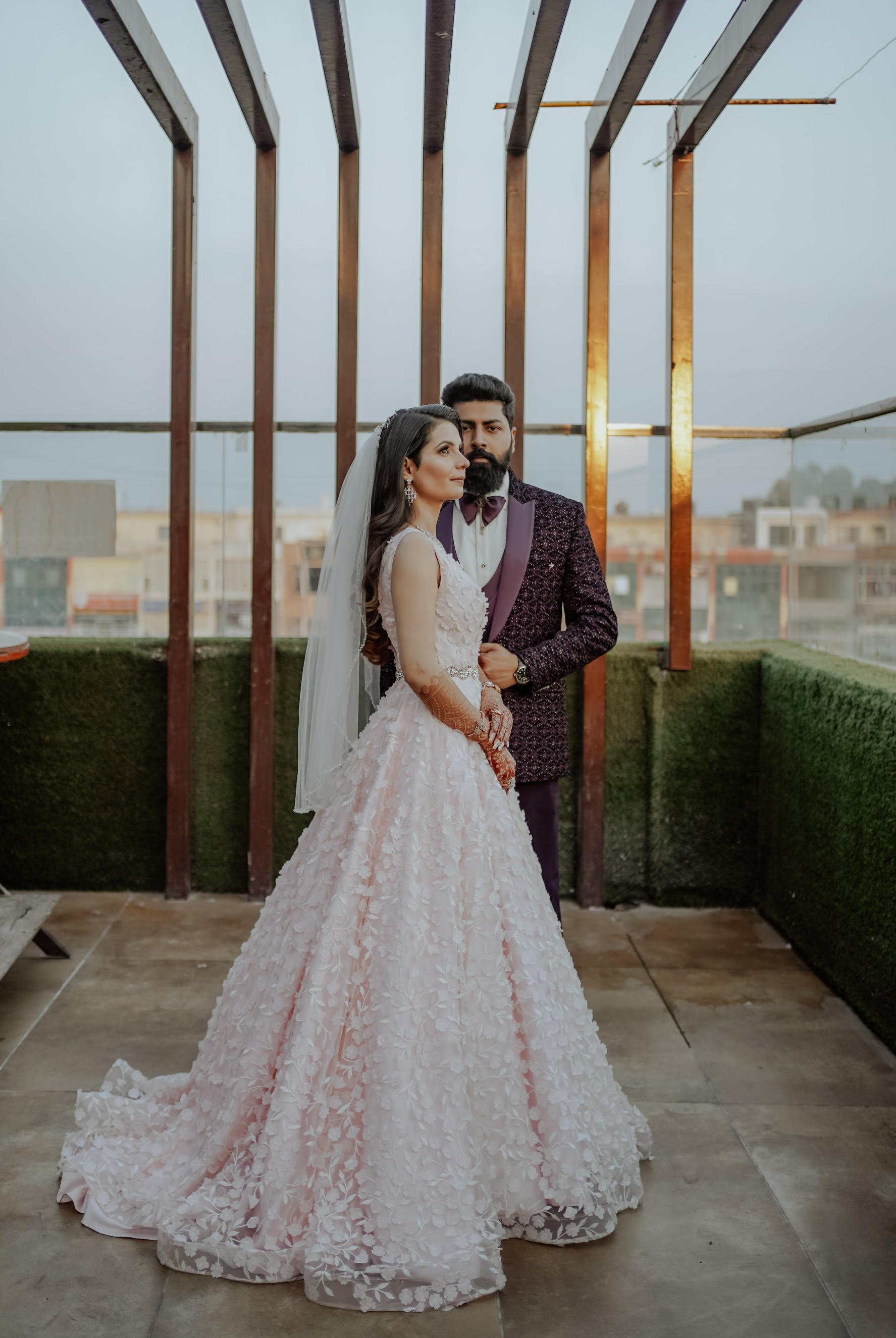 A bride in a pink gown and groom in a suit pose on a terrace with wooden beams and cityscape in the background.
