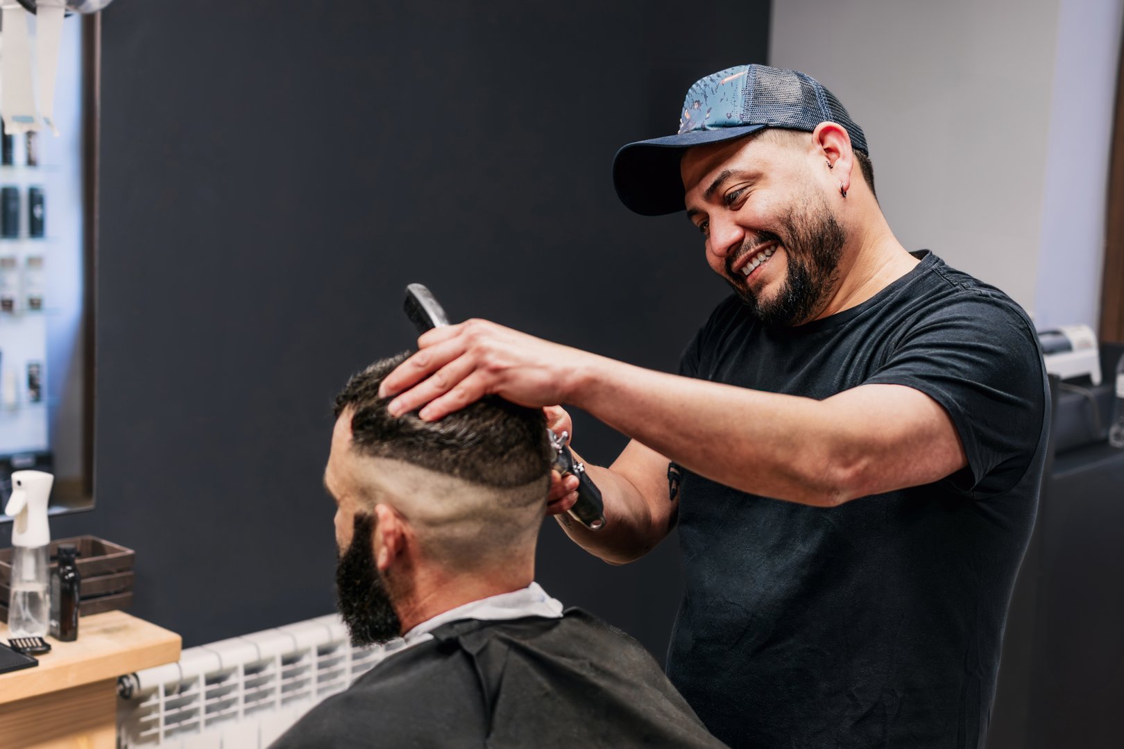 A Latin hairdresser cuts the hair of a man with a beard. The barber is wearing a baseball cap and is smiling and concentrating on his task.