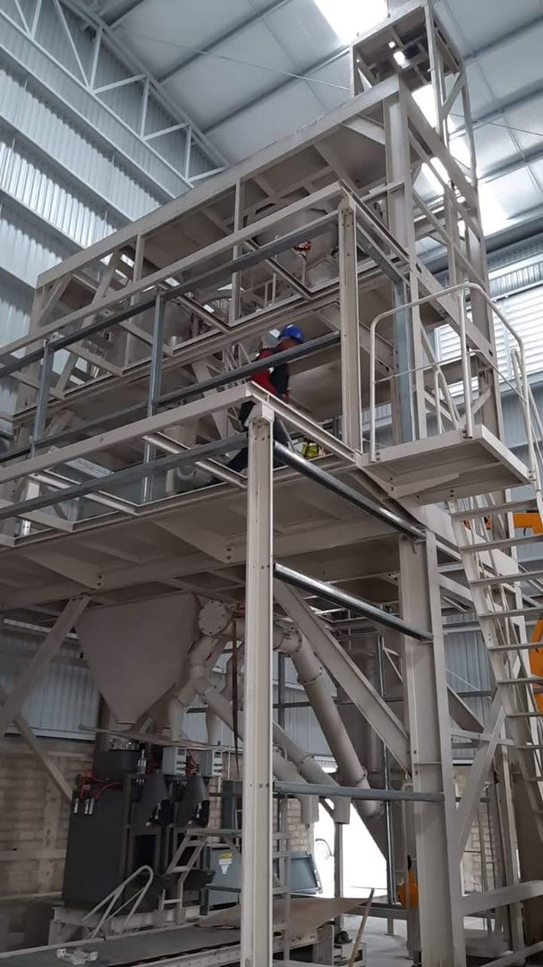 Worker in safety gear on industrial metal structure inside a factory, surrounded by steel beams and equipment.