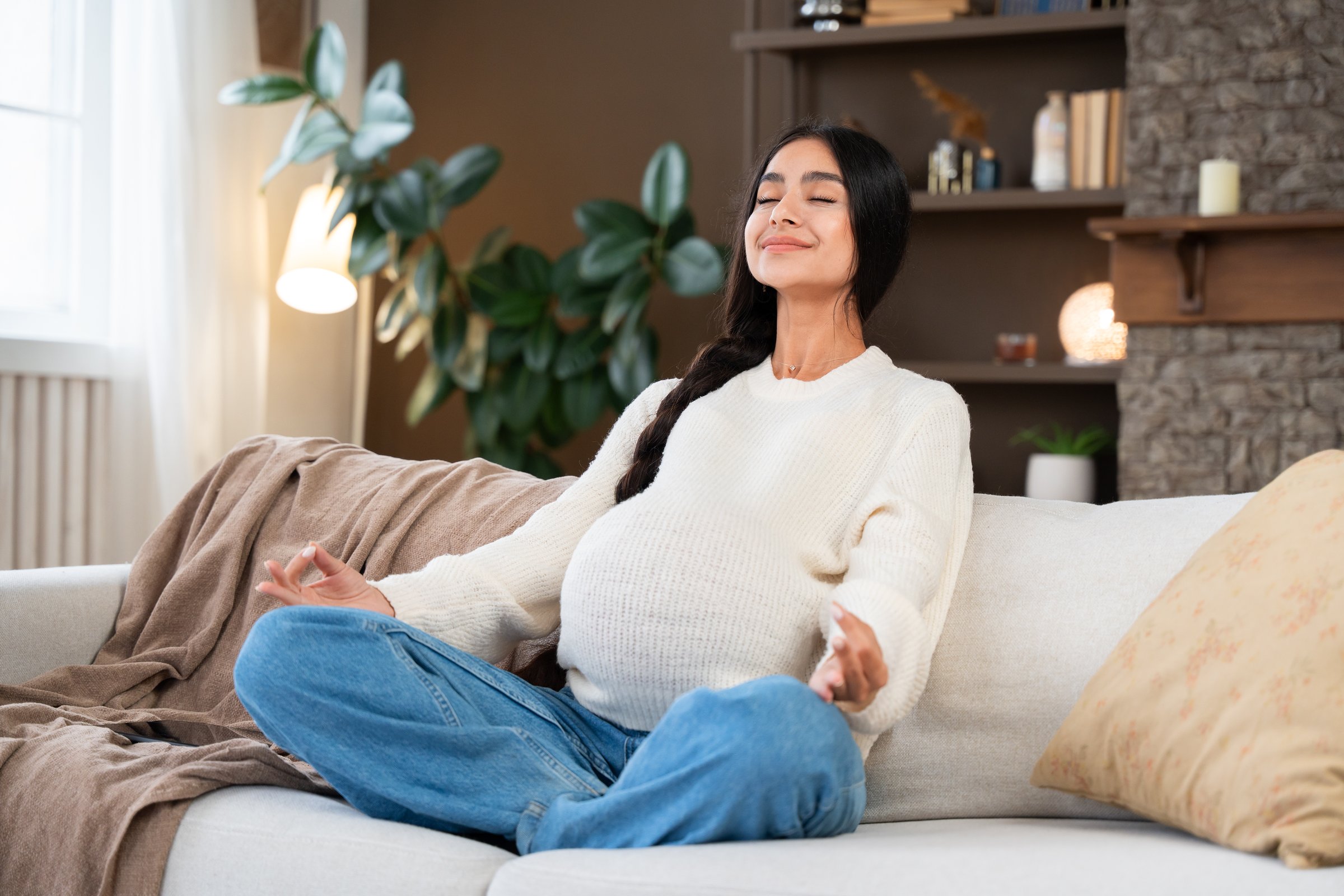 Pregnant Indian woman relaxing and meditating on the couch at home, engaging in mindful meditation to relax and ease her prenatal experience at home.