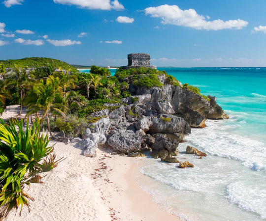 Scenic view of Tulum ruins on a rocky cliff overlooking turquoise ocean, surrounded by lush greenery and sandy beach.