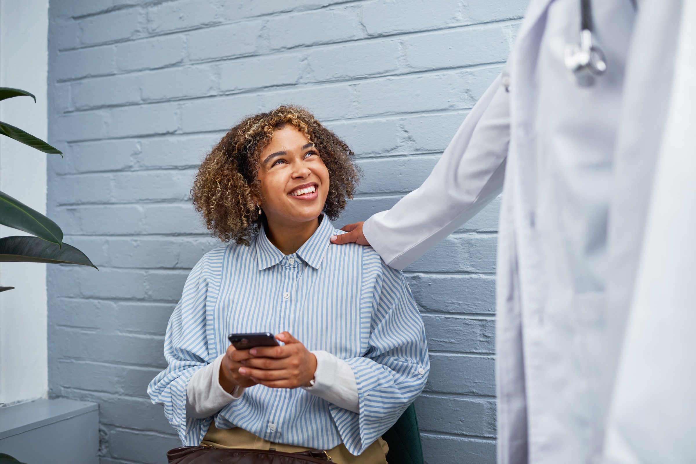 A cheerful young woman holding a smartphone sits in a waiting area. A doctor offers a comforting touch. The bright environment enhances the warm, compassionate interaction, symbolizing healthcare professionalism and understanding.