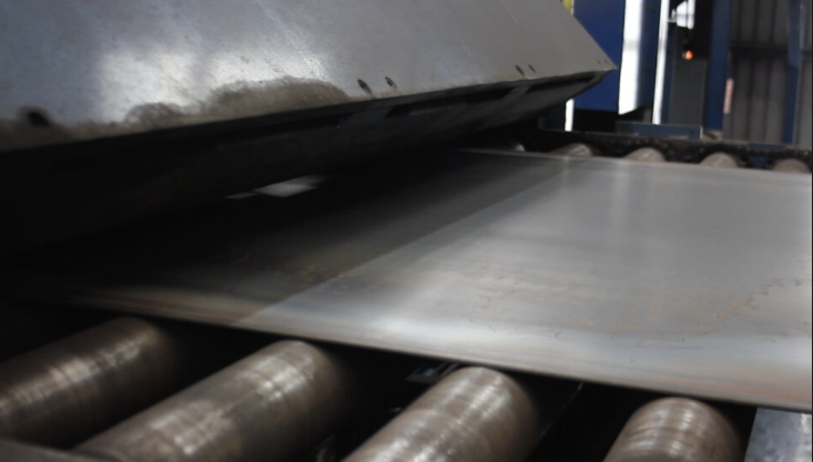 Sheet metal being processed in an industrial machine with rolling cylinders visible beneath.