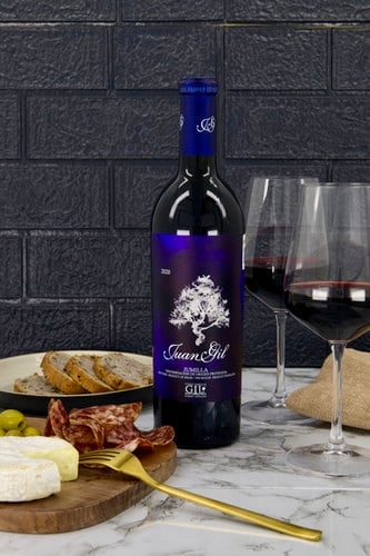 A bottle of red wine with two glasses, bread, cheese, and cured meat on a marble surface, against a dark tiled background.