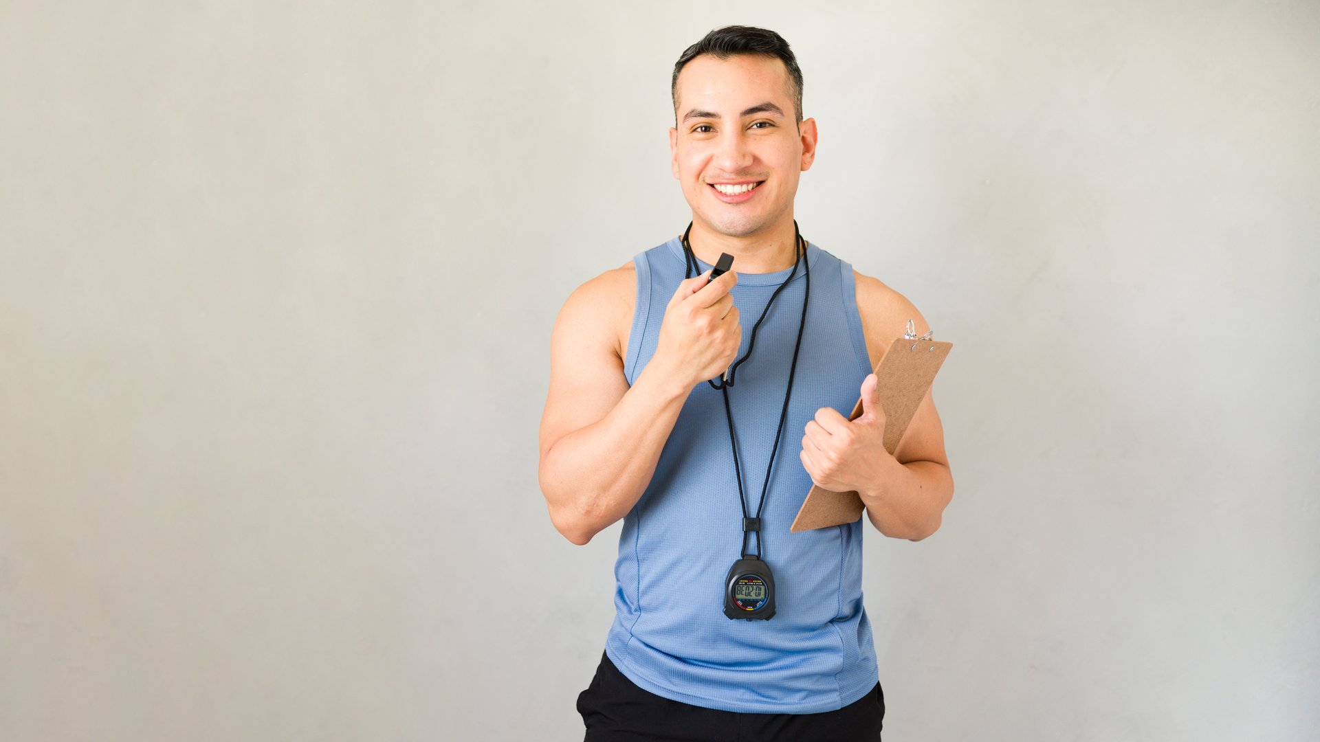 Cheerful hispanic male fitness coach is holding a clipboard and blowing a whistle while standing in front of a white background
