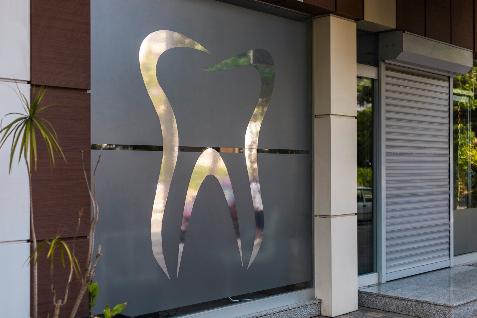 A modern dental clinic displays a stylish tooth logo on its glass window. The urban location has greenery visible nearby, highlighting the clinic's welcoming environment.