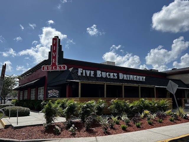 Exterior of Five Bucks Drinkery, a bar with a red facade and outdoor patio, under a partly cloudy sky.