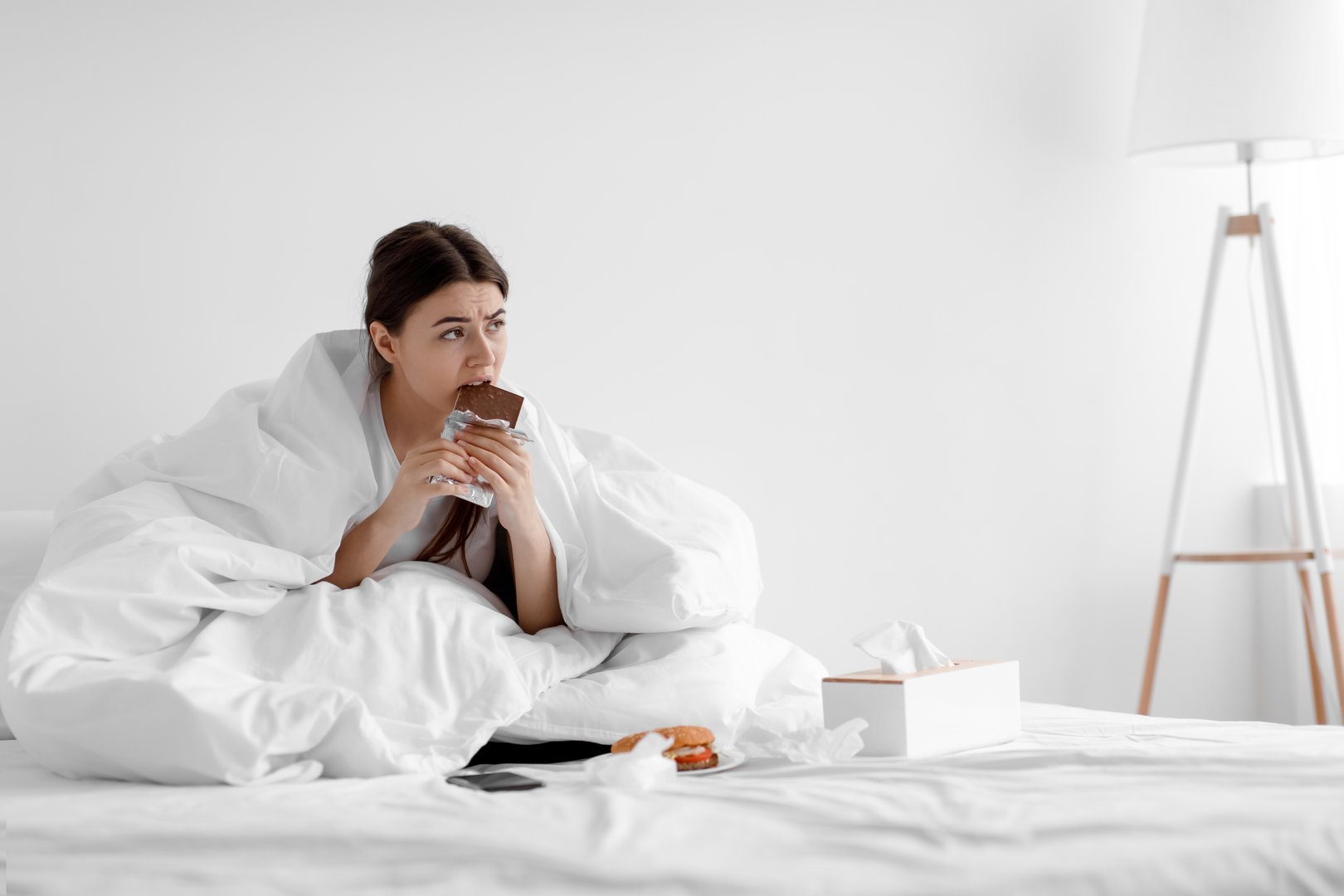 Upset hungry caucasian young woman sitting in bed at home and eating chocolate and junk food. Meal delivery service, eat addiction, disorder, psychological health, problems with appetite after breakup
