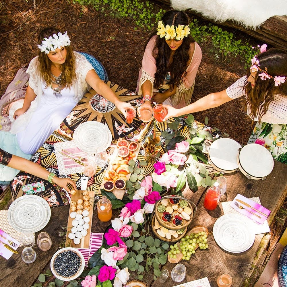 Four women having a picnic with food and drinks on a decorative table, wearing flower crowns in a woodland setting.