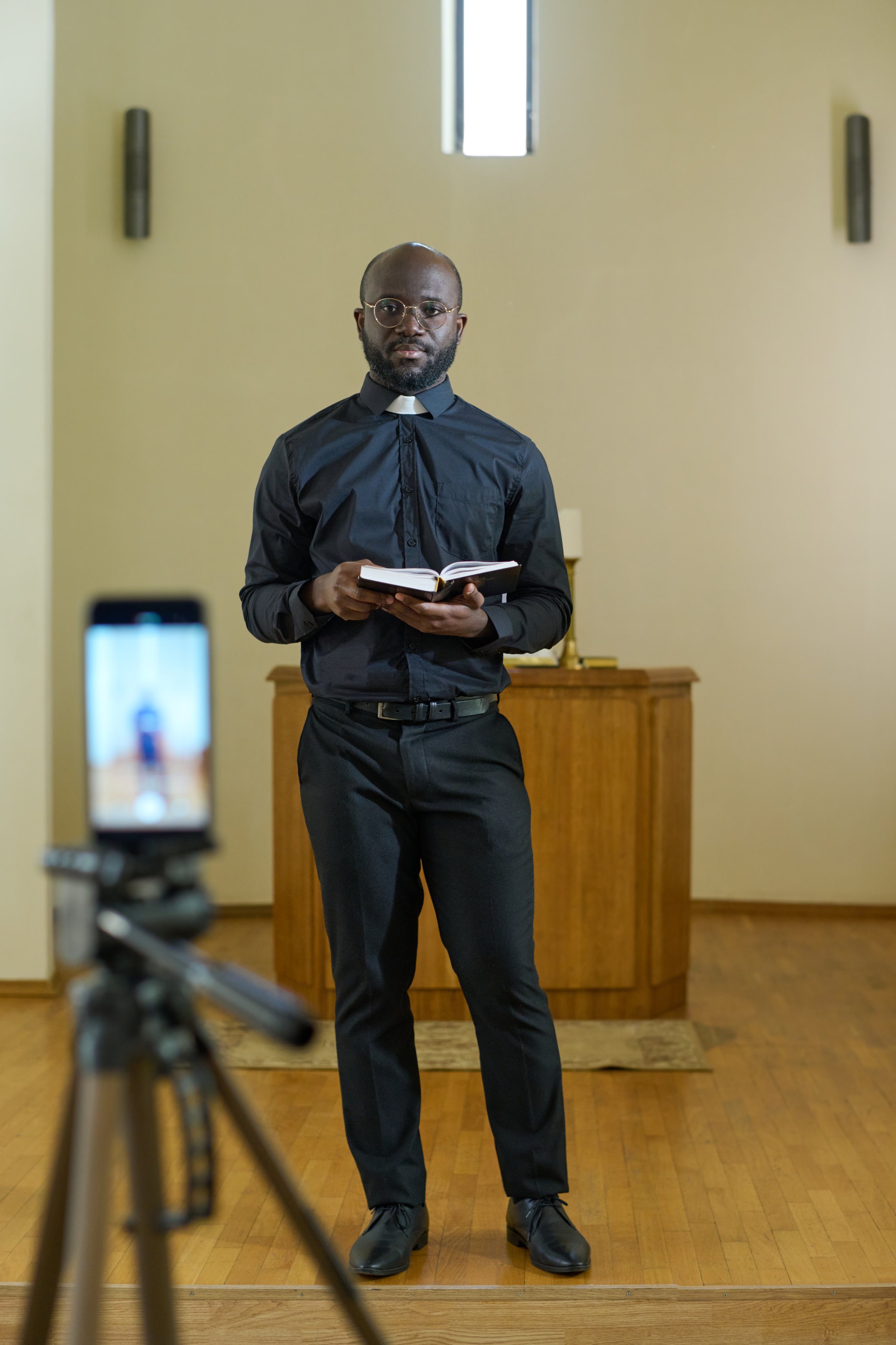 African American pastor in black pants and shirt with clerical collar holding open Bible while standing in front of smartphone camera