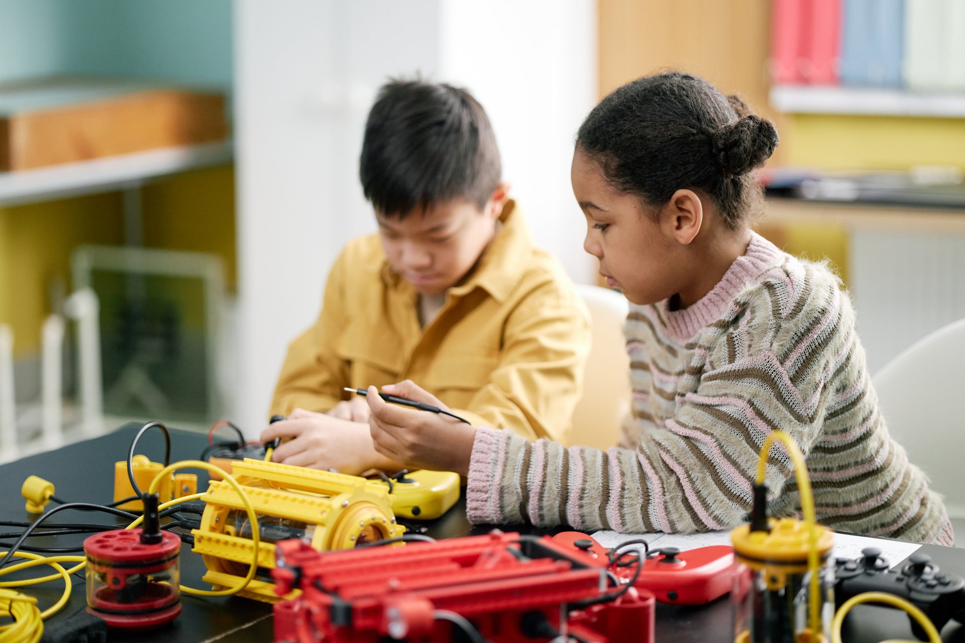 Students engaging with robotics kits on table in classroom, focusing on building and programming. Children appear attentive and involved in project, representing hands-on learning environment