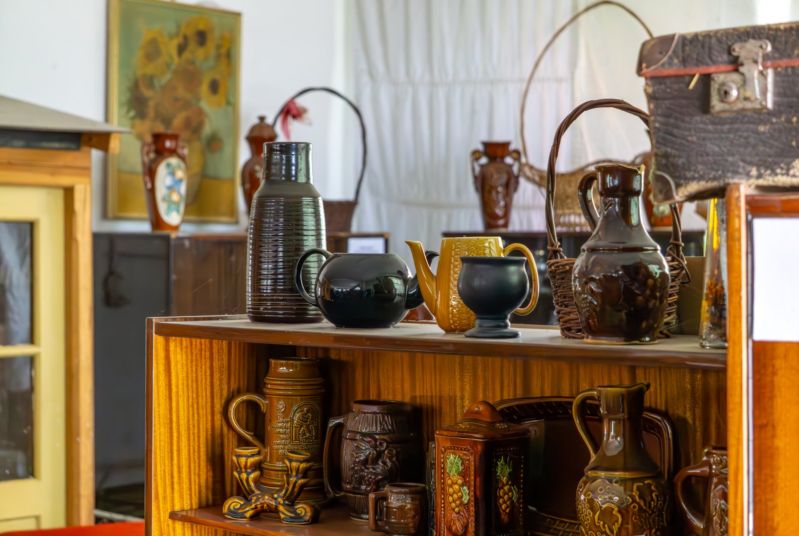 Wooden shelf with a collection of vintage ceramic jugs, mugs, and teapots, set against an interior with a painting and an old suitcase