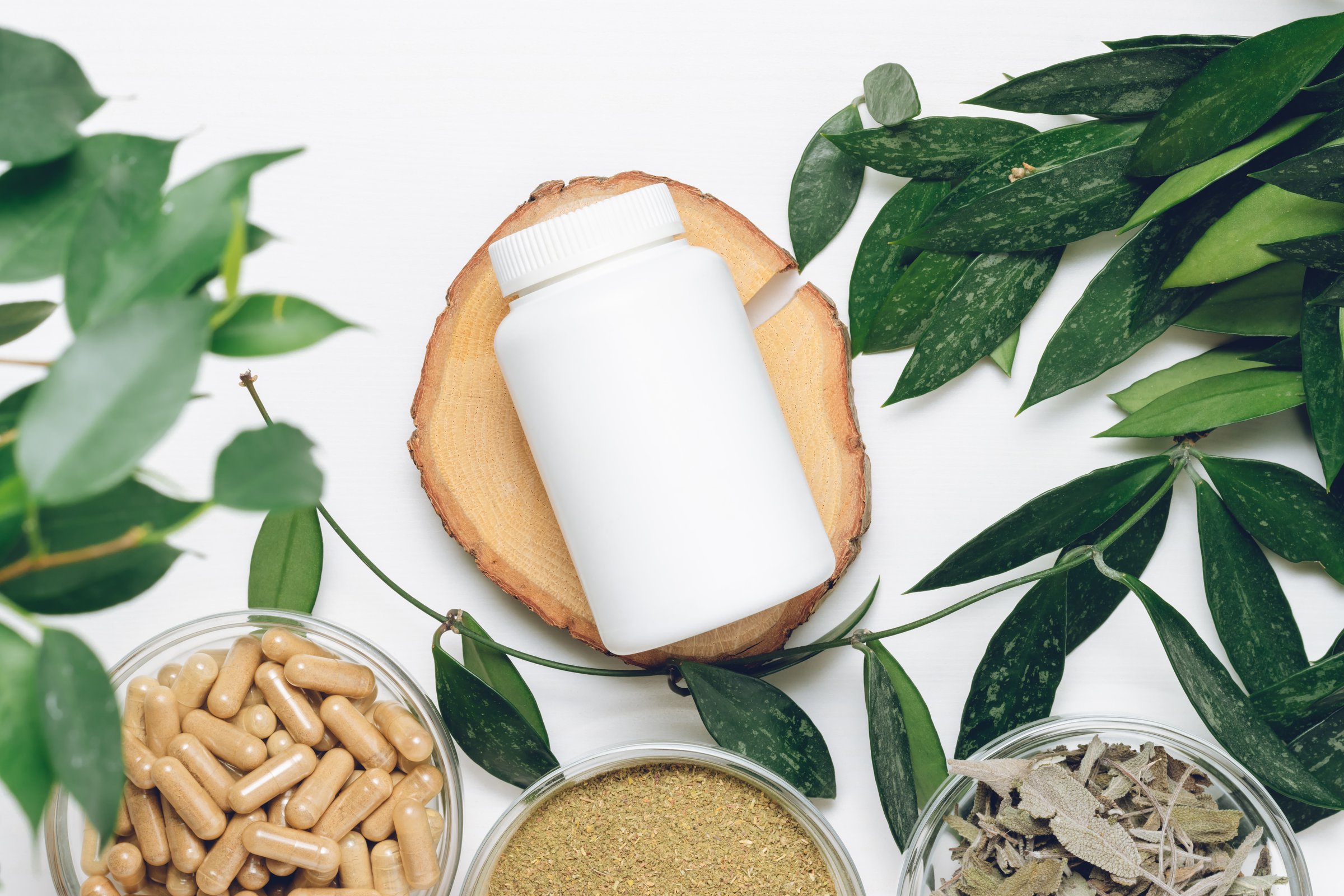 White bottle for supplement on wooden podium with green leaves, herbal pills and ingredients, top view. Organic vitamins for health