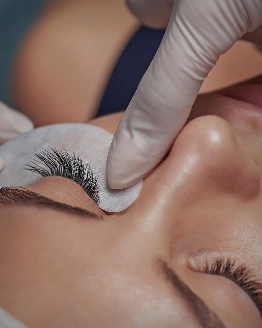 Close-up of a woman receiving an eyelash extension treatment with a technicians gloved hand adjusting under-eye pads.