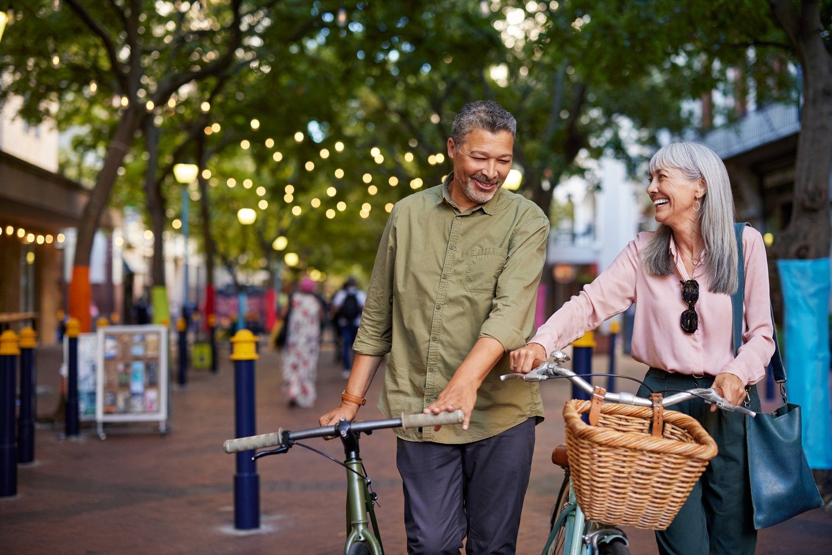 Smiling old couple walking with bicycles in the street with copy space. Happy senior multiethnic man and beautiful mature woman enjoying a bike ride together in town. Multiethnic elderly couple on a romantic evening walk laughing while pushing their bike in a pedestrian area.