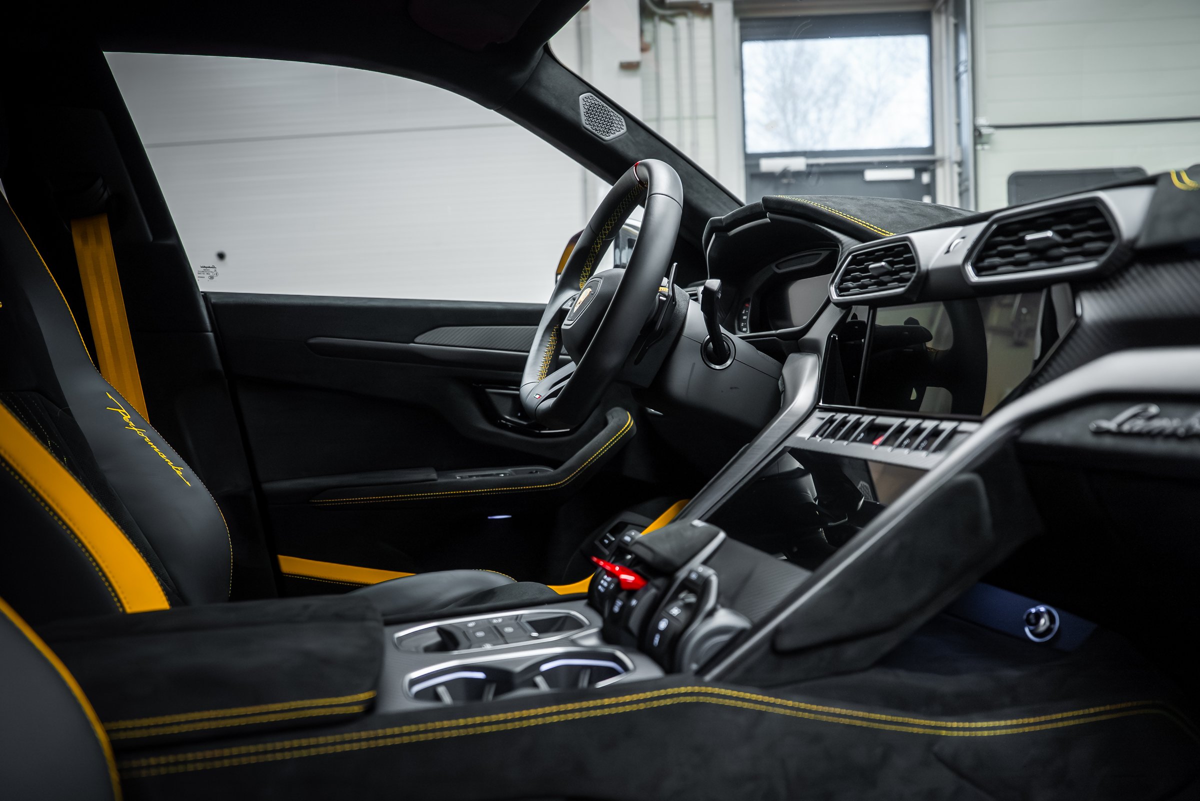 Driver focused interior shows bucket seats with yellow accents, carbon trim, hexagonal vents, dual screens, and fighter jet style switch in a garage with cool diffused light.