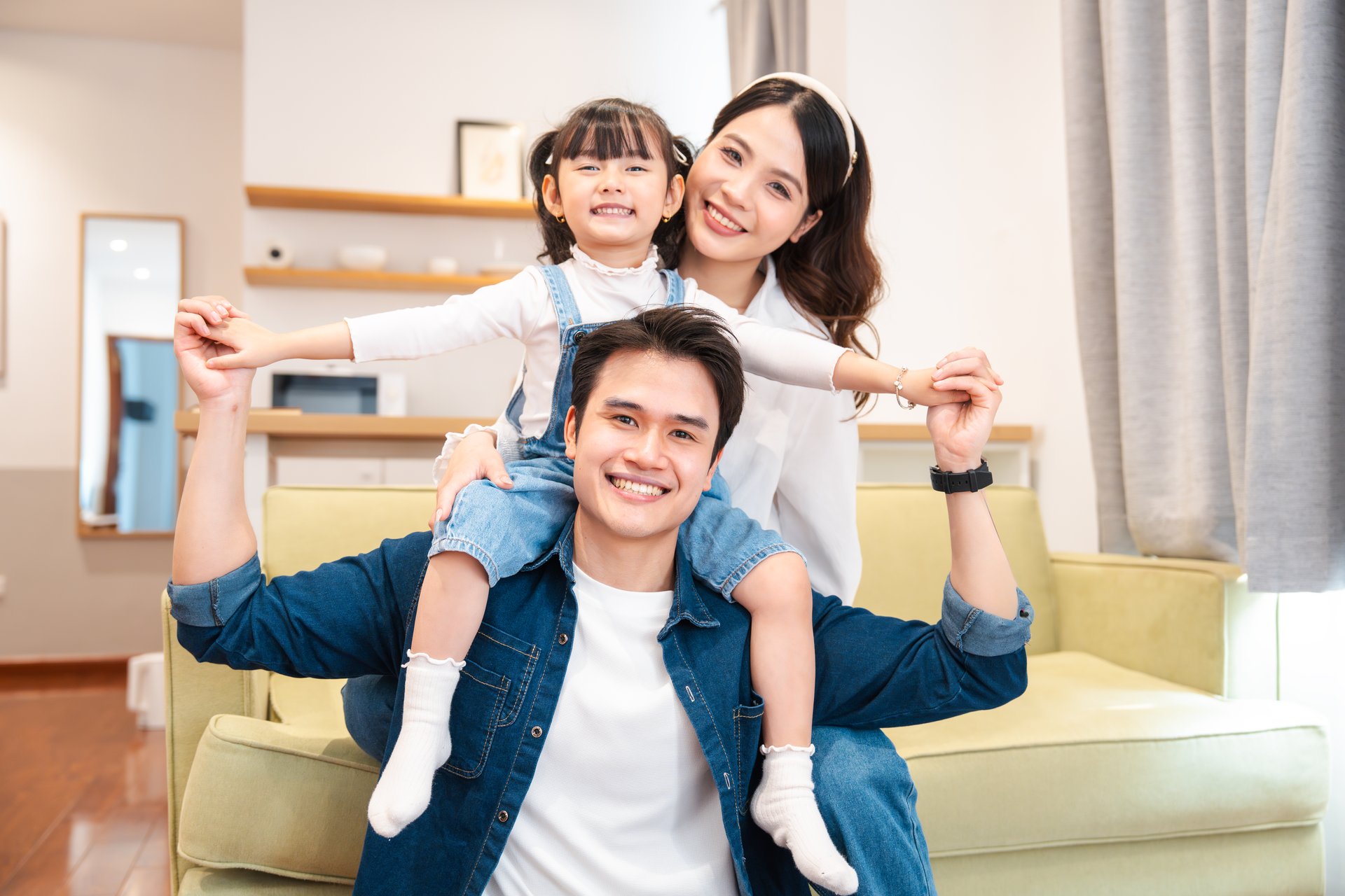 A happy family of three poses indoors The father carries his daughter on his shoulders as mother smiles