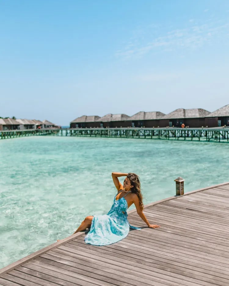 Woman in a blue dress sitting on a wooden deck by turquoise water, with overwater bungalows in the background.