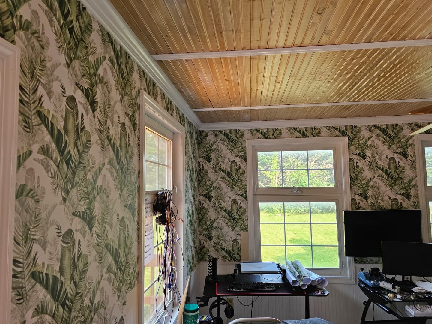 Room with tropical leaf wallpaper, wooden ceiling, two windows, a desk, computer, and a scenic view outside.