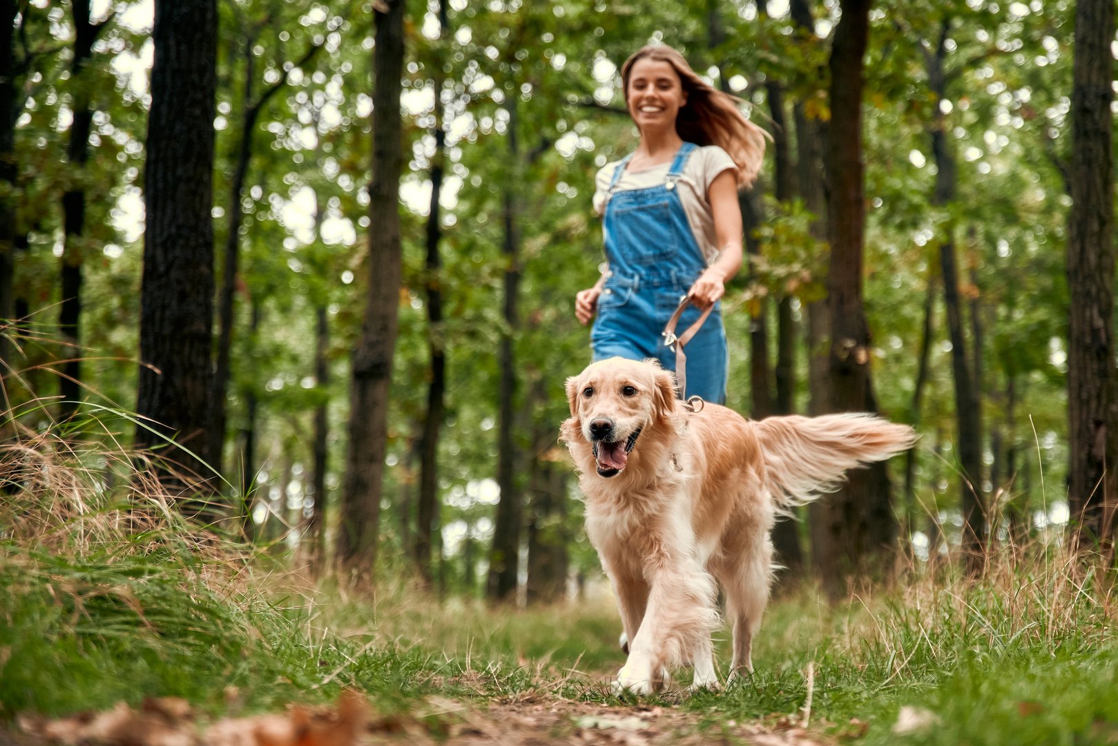 A joyful woman in comfortable overalls walks through a lush green forest with her playful golden retriever, both enjoying the wonders of nature and the delightful companionship of her pet