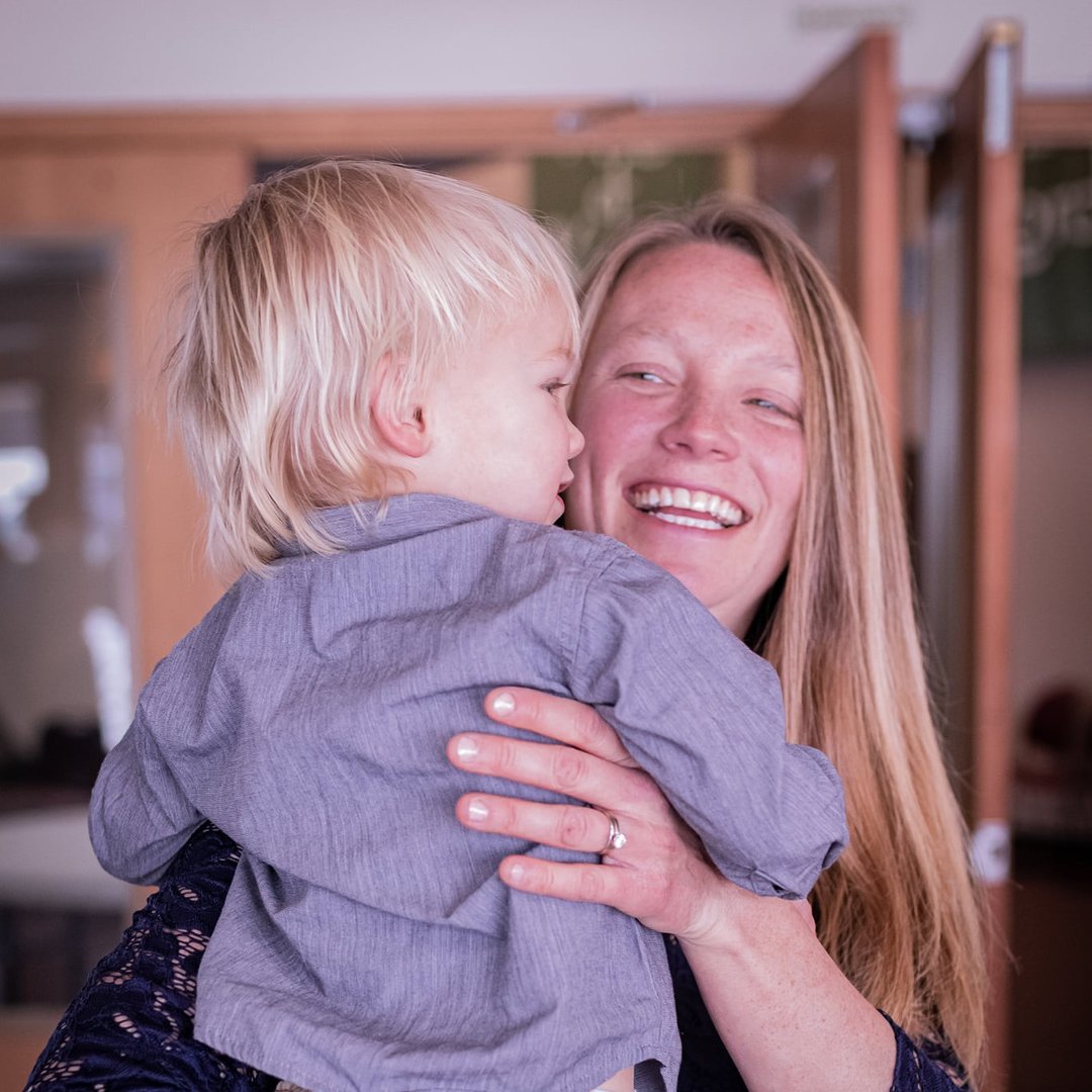 A smiling woman holding a young child indoors, both with light-colored hair.