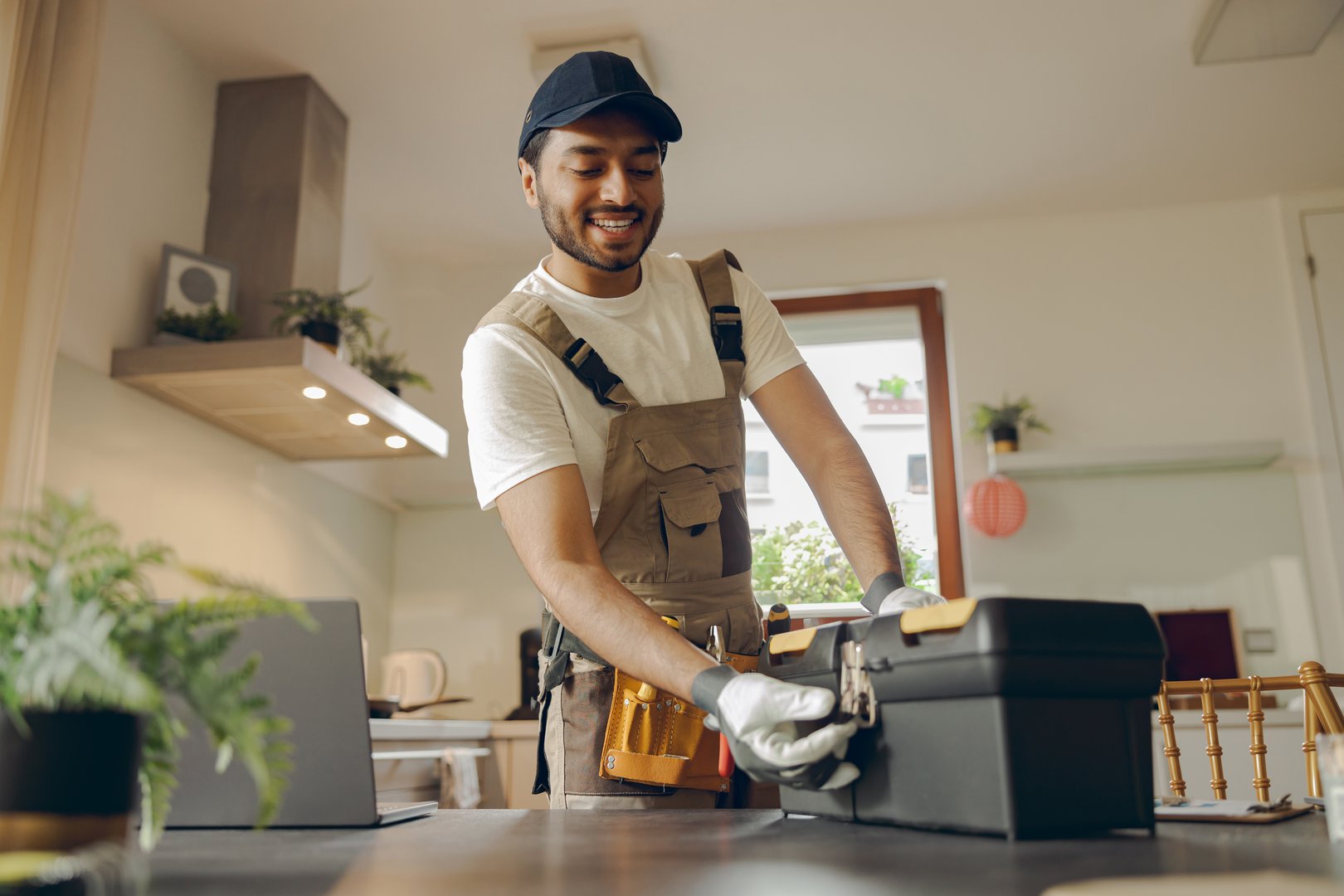 Positive repairman in uniform standing on home kitchen and opening his tool bag
