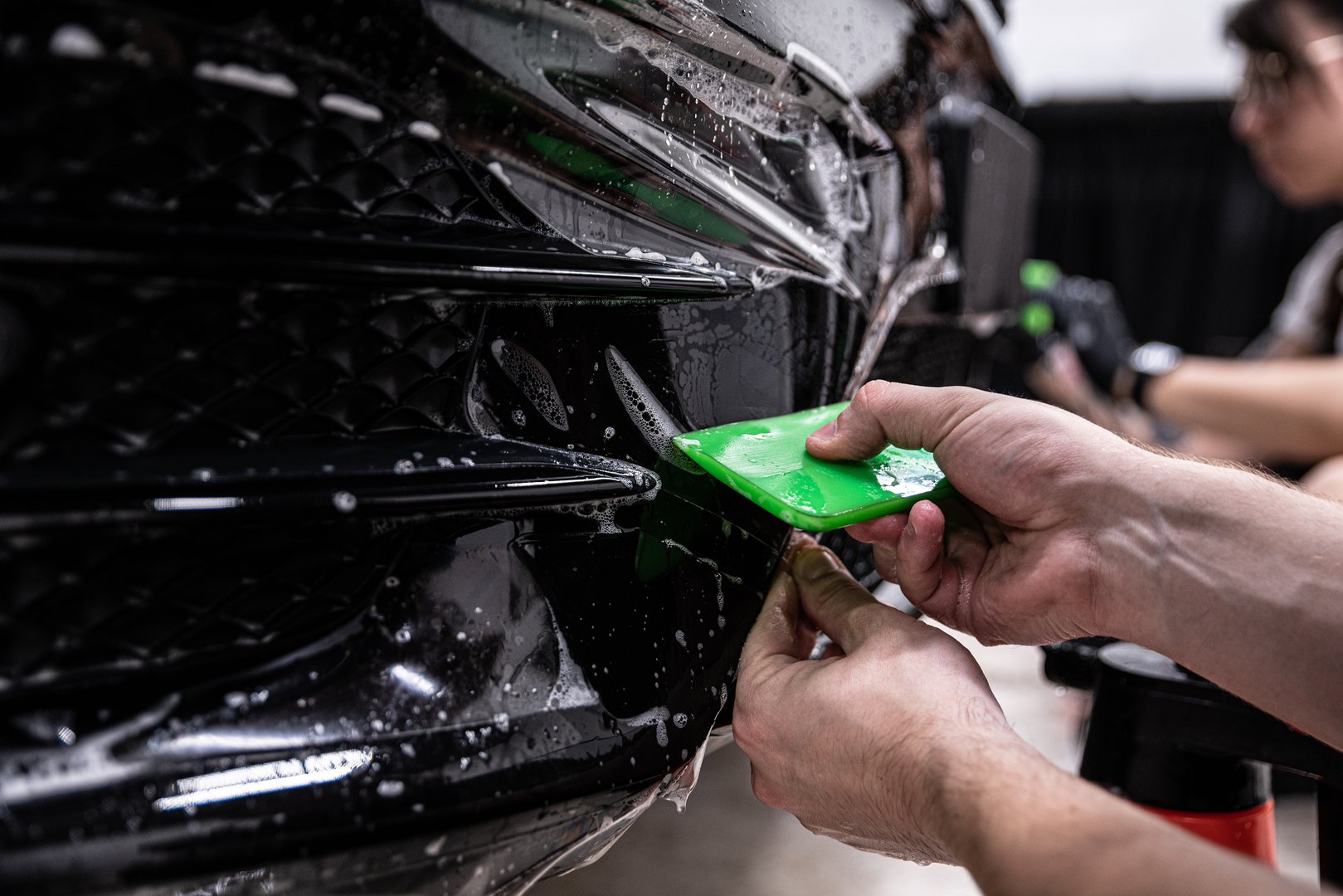 Car detailing studio employee, a wrapper, is protecting the bumper of a black car with a colorless protective PPF film. The PPF film protects the paint from scratches and damage.