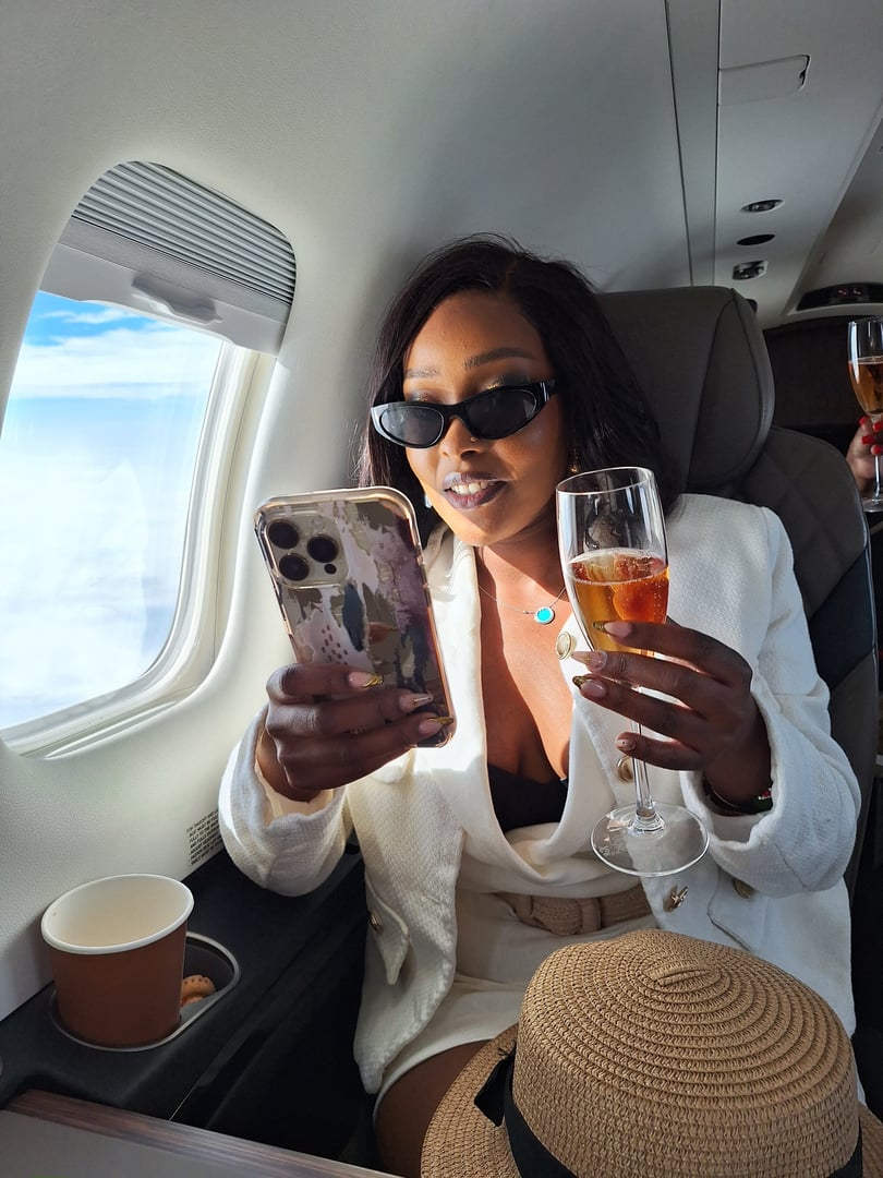 Woman in white outfit holding a smartphone and champagne glass, seated in an airplane, wearing sunglasses.