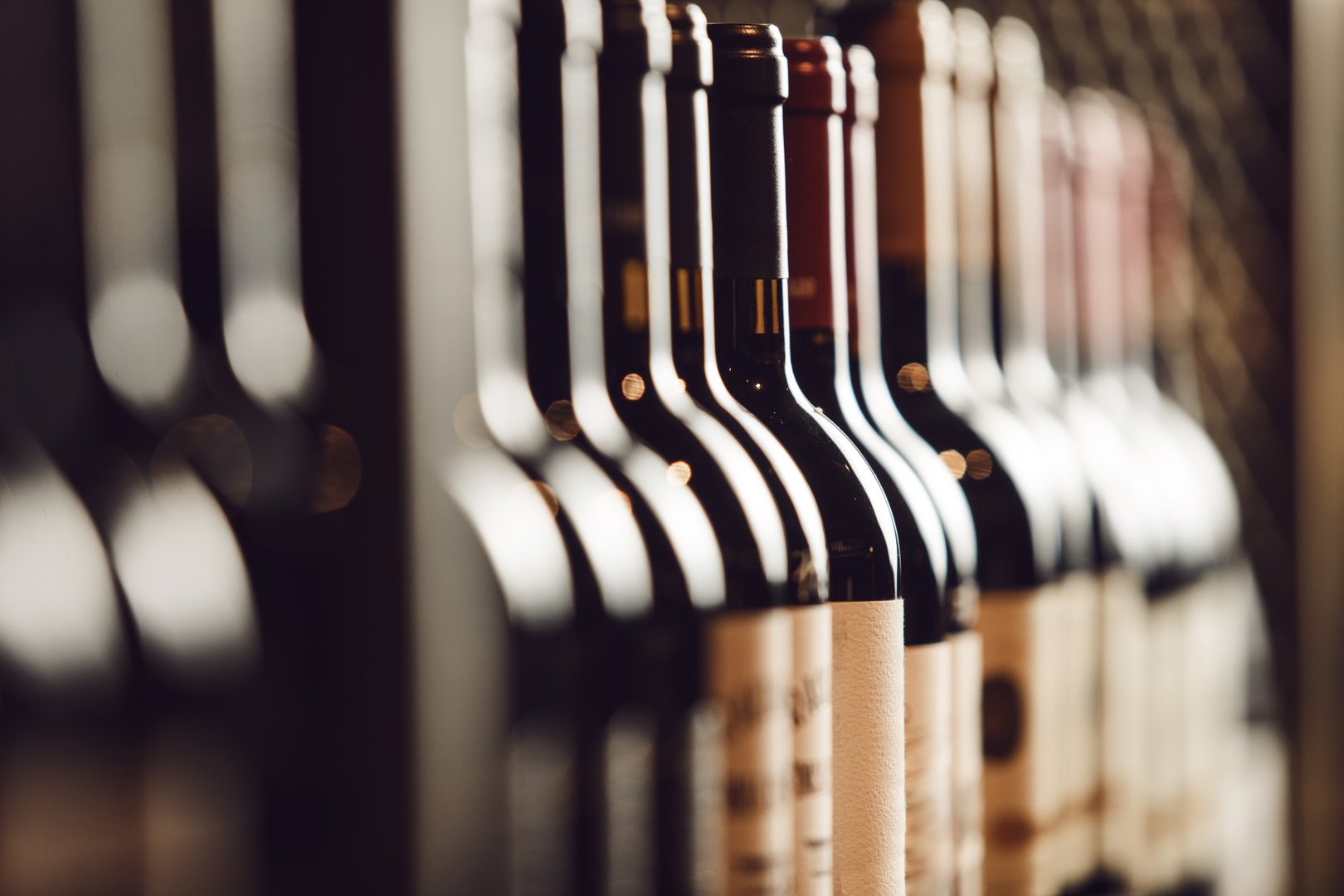 Rows of wine bottles with focus on the nearest bottles with labels facing forward are neatly arranged on wooden shelf in cellar, showcasing variety of elite alcohol options for connoisseurs.