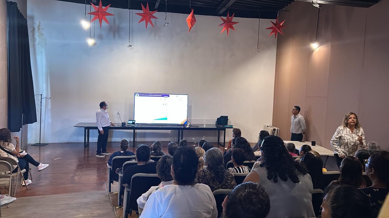 Group of people attending a presentation in a room with red star decorations on the ceiling. Presenter at the front near a screen.