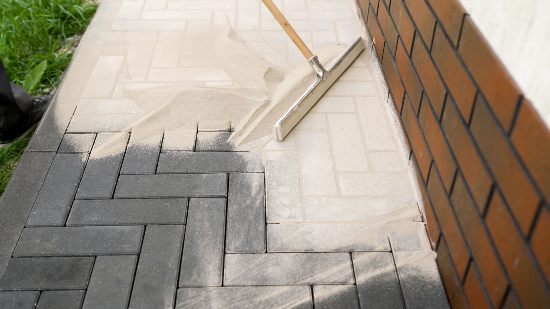 Construction worker uses a squeegee to spread sand over paving stones near a brick wall, creating a stable and level surface