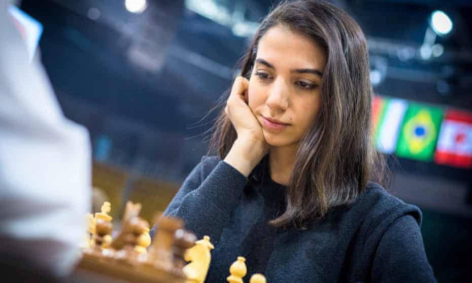 A woman intently playing chess, resting her chin on her hand. National flags are visible in the blurred background.