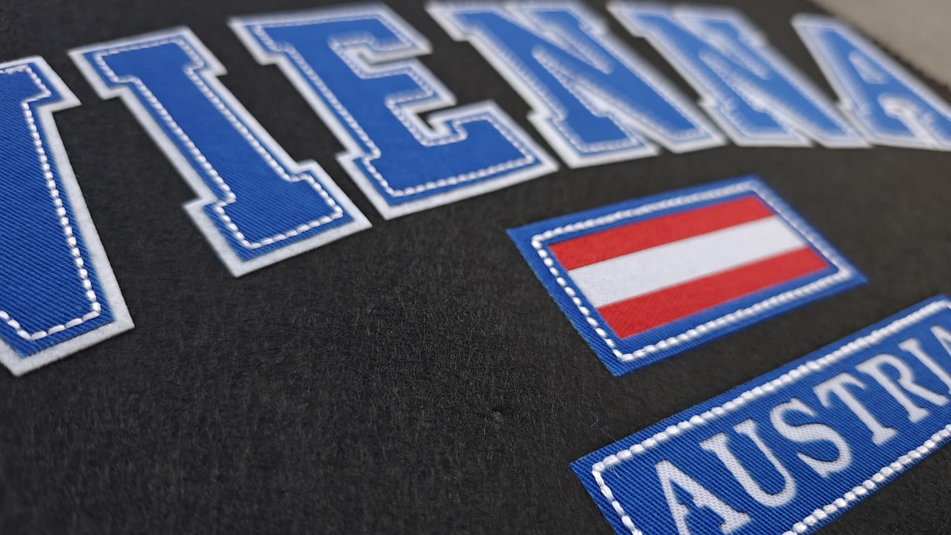 Close-up of a black fabric with blue stitched letters spelling Vienna above an Austrian flag and Austria in white.
