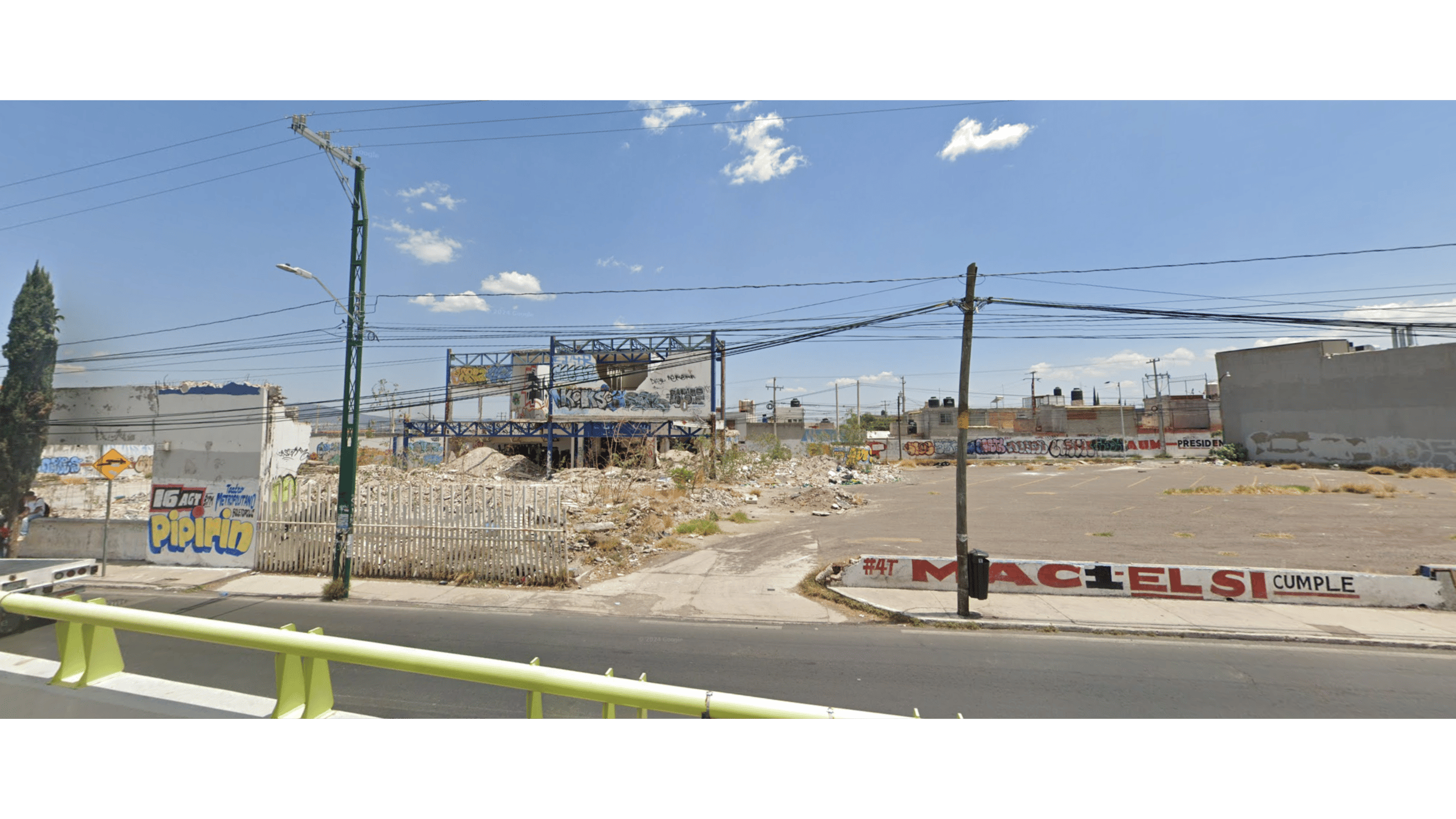 Vacant lot with rubble, surrounded by graffiti-covered walls and a few buildings, under a partly cloudy sky.