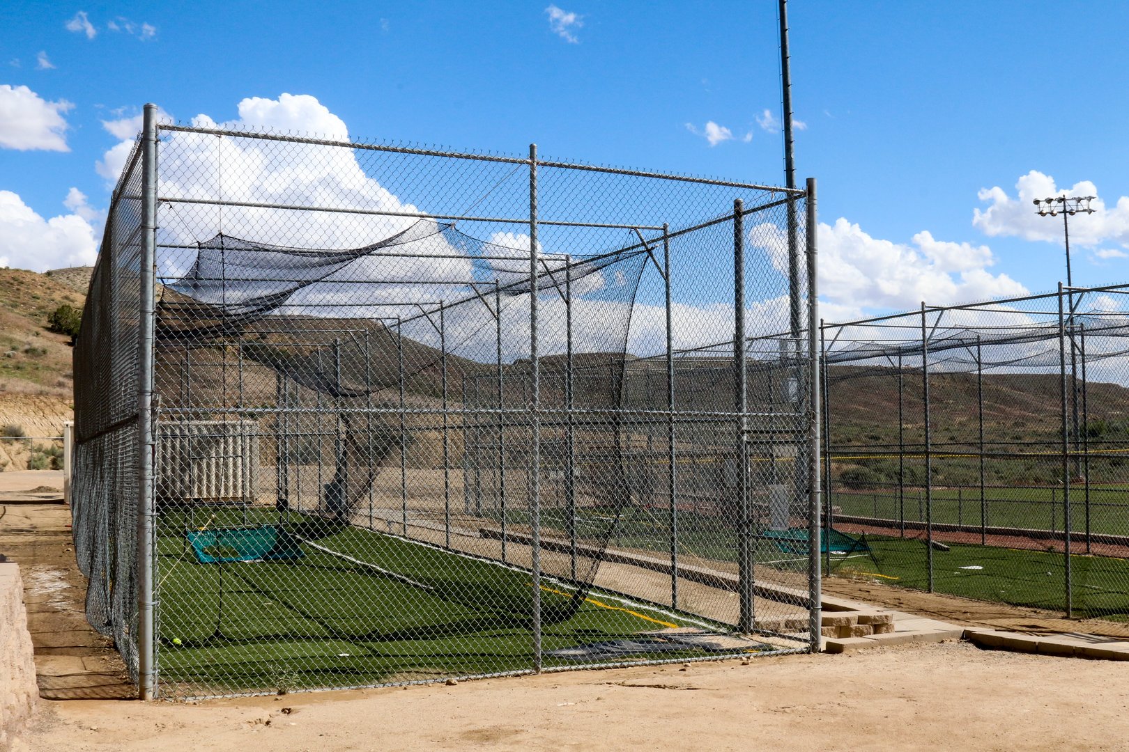 Image of a fully enclosed batting cage.  Batting cages are used to practice hitting the ball in softball and baseball.  The sky overhead is blue with puffy cumulous clouds.  Inside the cage is green, fake turf.  The photo was taken in New Mexico.
