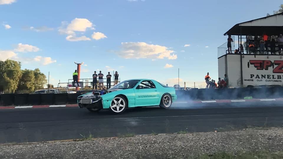 A turquoise car drifting on a race track, creating smoke, with spectators and a building in the background.
