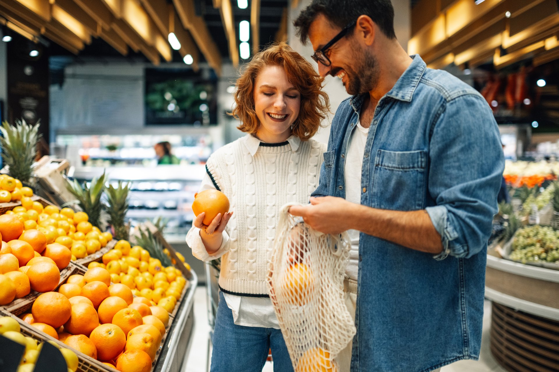 Young smiling couple buying fresh oranges at supermarket, holding reusable mesh bag, promoting sustainable and eco friendly lifestyle, choosing healthy food